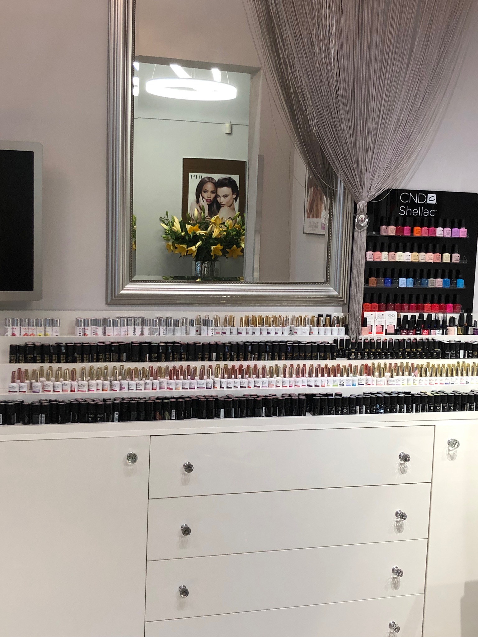 Salon kosmetyczny: biała komoda z rzędami lakierów do paznokci w różnych kolorach, lustro z kwiatami i plakatami, oraz ekspozytor CND Shellac.