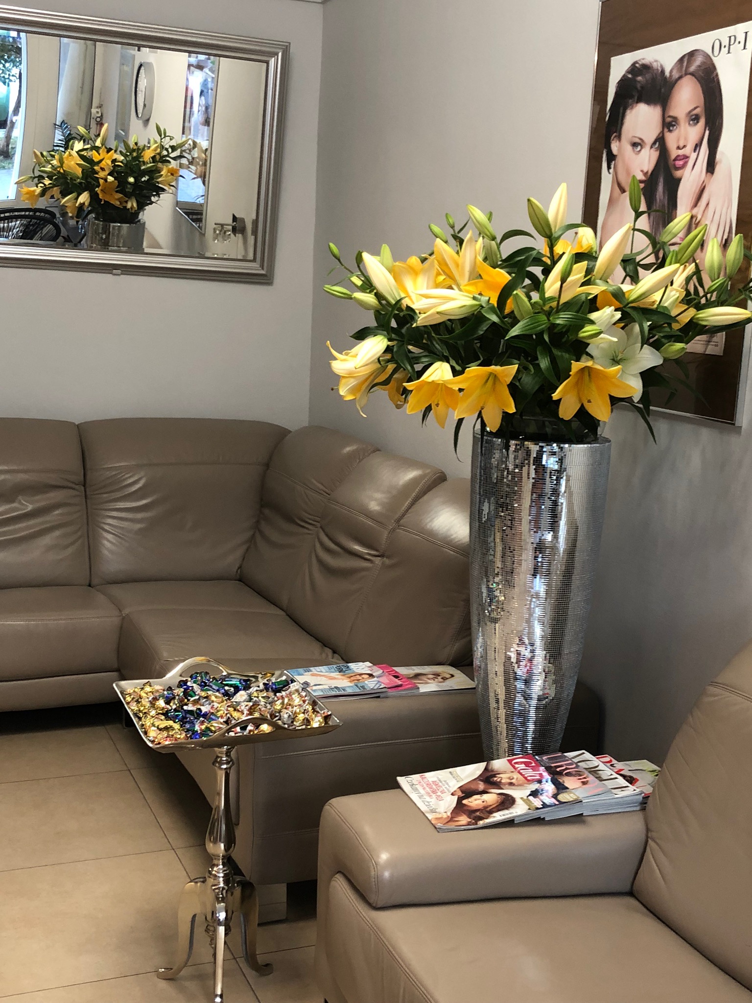Wnętrze salonu kosmetycznego z beżową sofą, srebrnym wazonem z żółtymi liliami, stolikiem z cukierkami i stosem magazynów, plakat reklamowy OPI na ścianie.