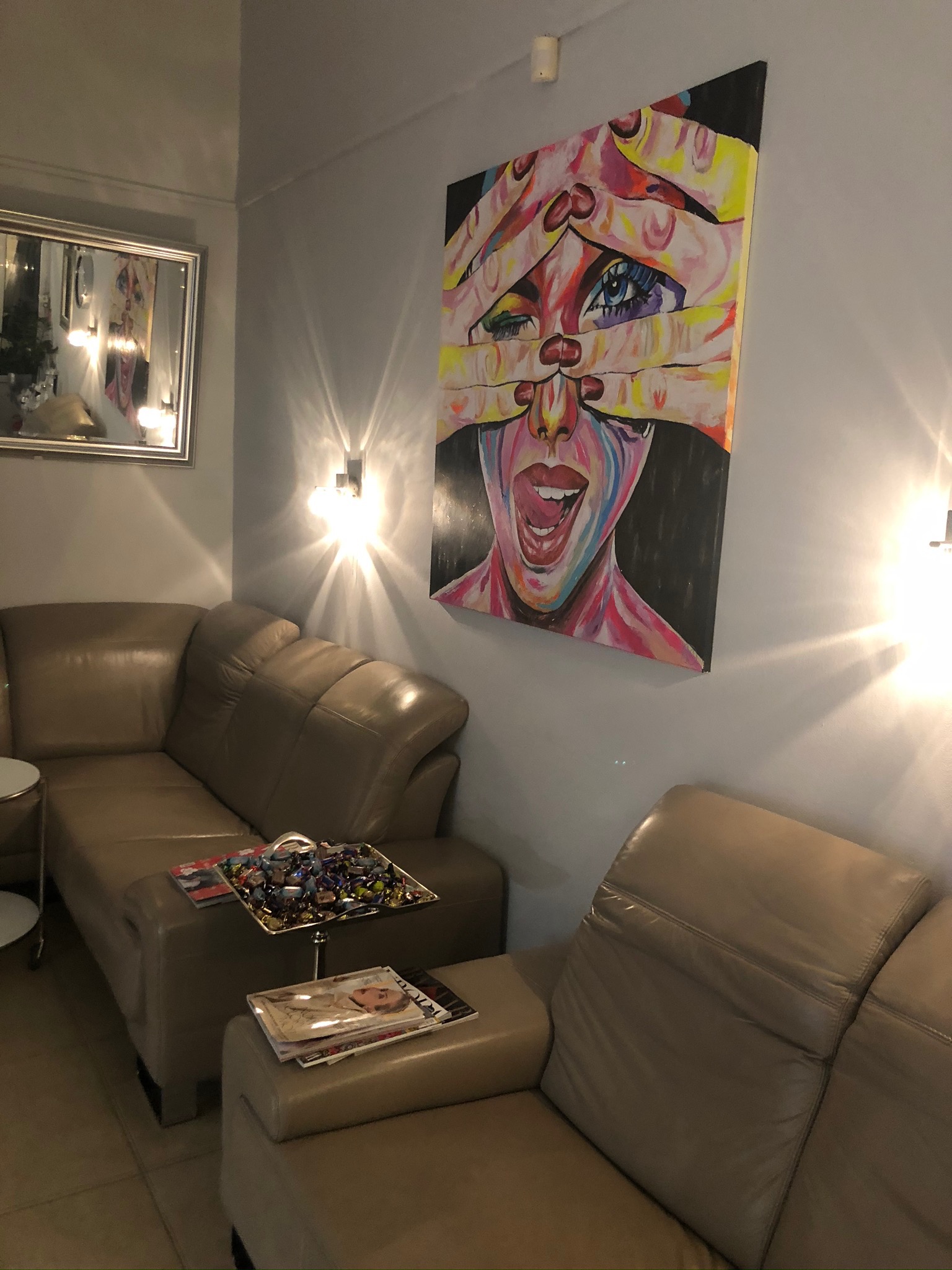 Poczekalnia salonu kosmetycznego z beżowymi kanapami, stolikiem z czasopismami i cukierkami, oświetlona kinkietami, z obrazem kobiety zakrywającej oczy na ścianie.