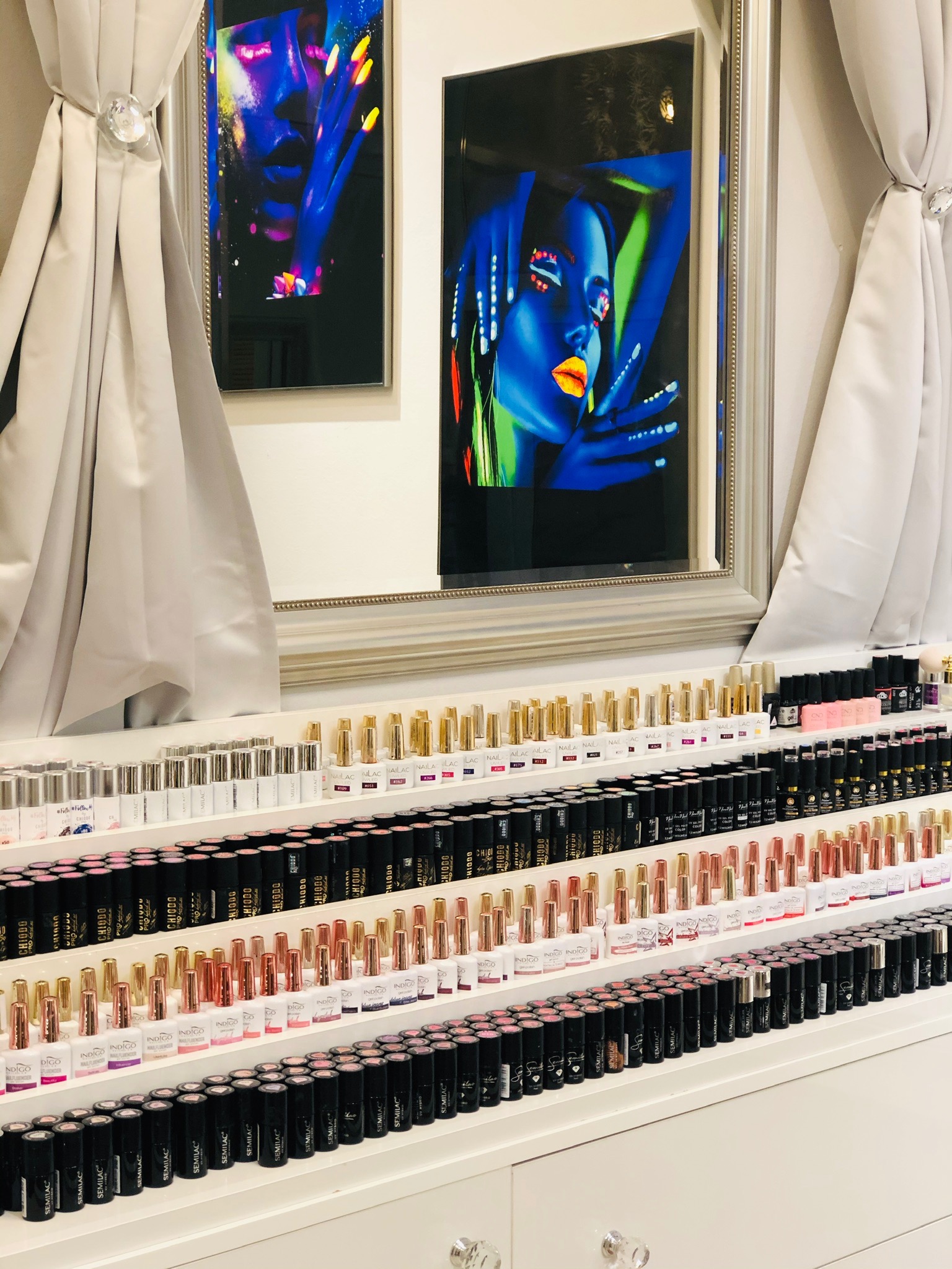 Szeroki kadr na ekspozycję lakierów do paznokci w salonie kosmetycznym, z podświetlonymi obrazami na ścianie i drapowanymi zasłonami po bokach.