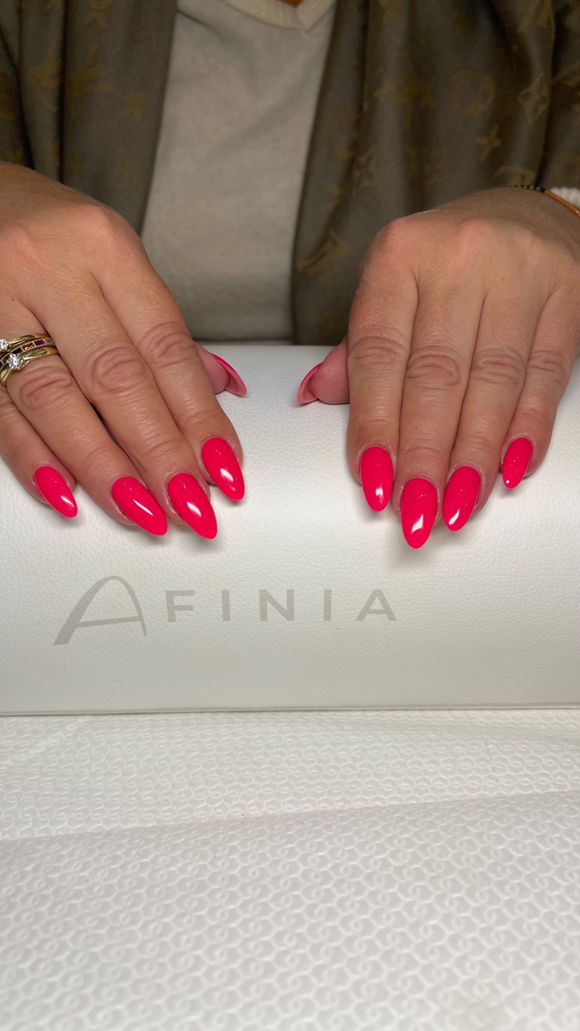 Ręce kobiety z jaskrawo różowymi, migdałowymi paznokciami na białym podłożu z logo Afinia, widoczny pierścionek z trzema kamieniami na palcu.