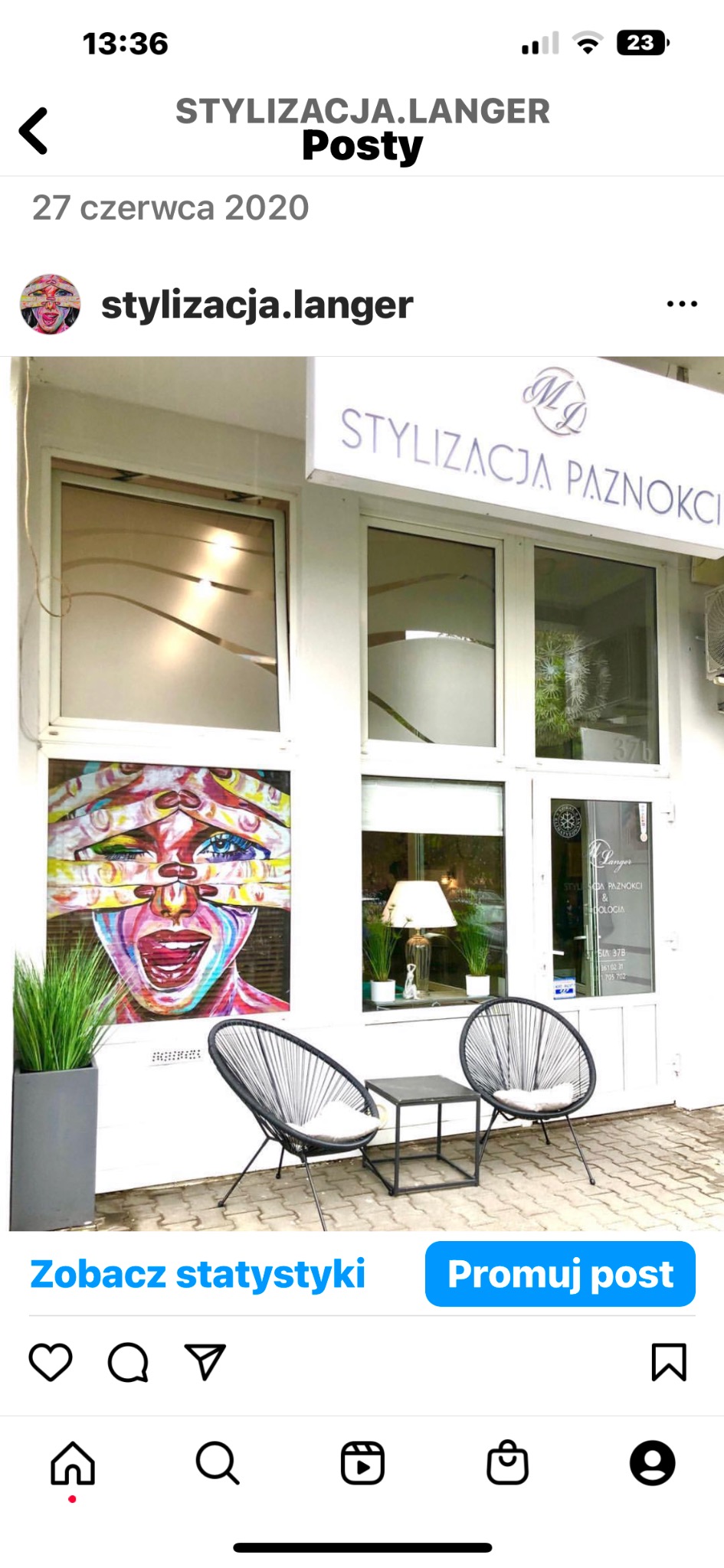 Wejście do salonu stylizacji paznokci z napisem 'Stylizacja Paznokci' nad drzwiami, ozdobione grafiką na oknie, z czarnymi krzesłami i stolikiem na zewnątrz.