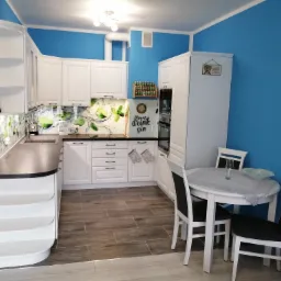 Jasna kuchnia z białymi meblami, drewnopodobnymi płytkami na podłodze i turkusowymi ścianami. Na blacie kuchennym dekoracyjny panel z motywem limonki, a nad nim półka z przyprawami. W rogu stoi...