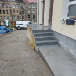 Zewnętrzne schody z szarego granitu prowadzące do budynku z beżową elewacją, betonowy podjazd dla wózków, w tle budynki mieszkalne i biały samochód dostawczy.