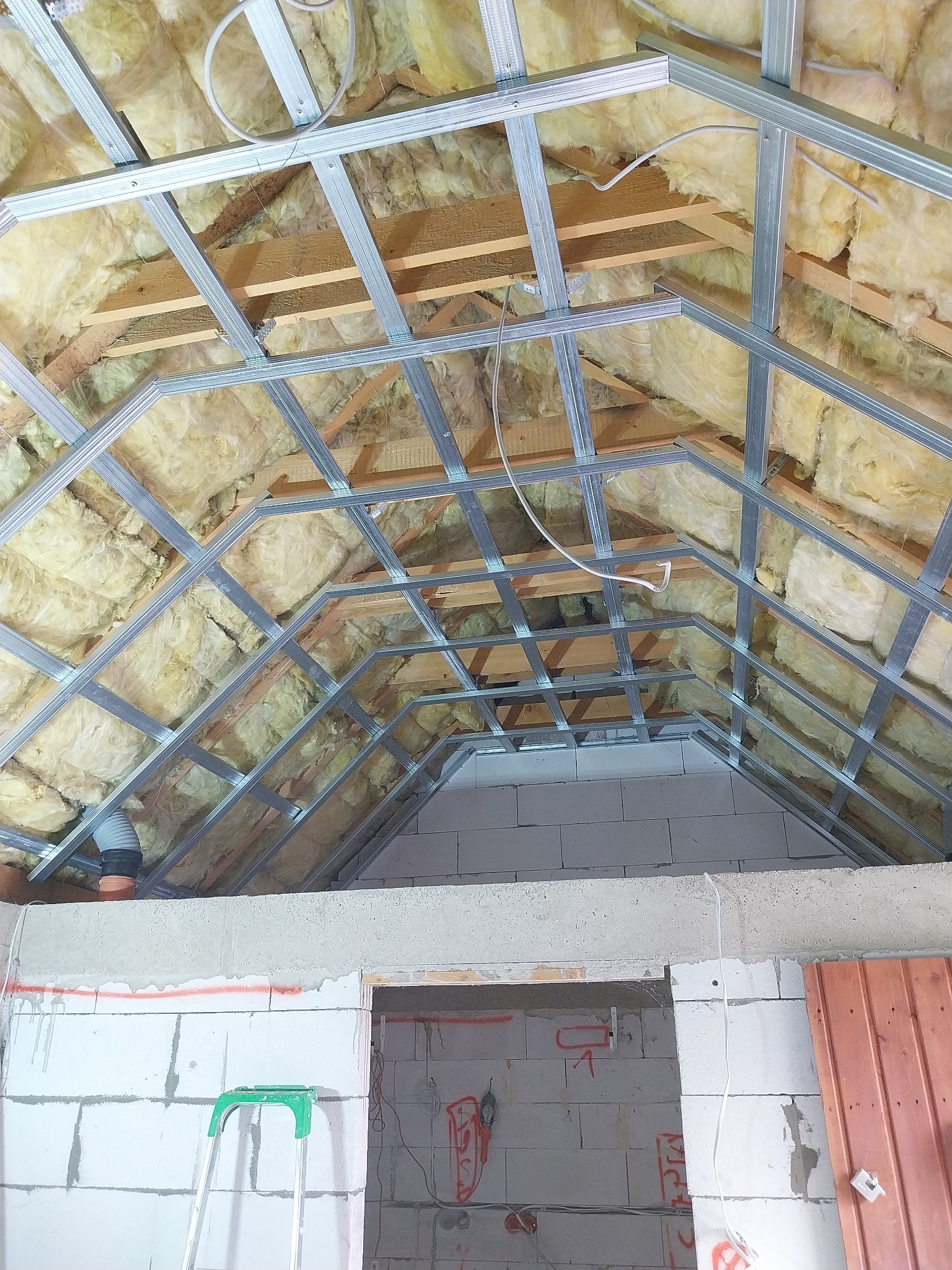 Montaż metalowej konstrukcji sufitu podwieszanego z izolacją z wełny mineralnej na poddaszu w Zgorzelcu, widoczny fragment ściany z betonu komórkowego i drabina.