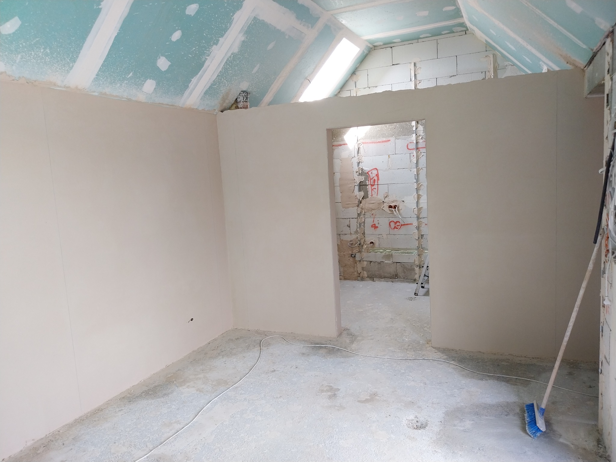 Wnętrze w trakcie prac wykończeniowych: ściany po gładzi, sufit z płyt gipsowo-kartonowych, widoczne instalacje w surowej ścianie i szczotka oparta o ścianę.