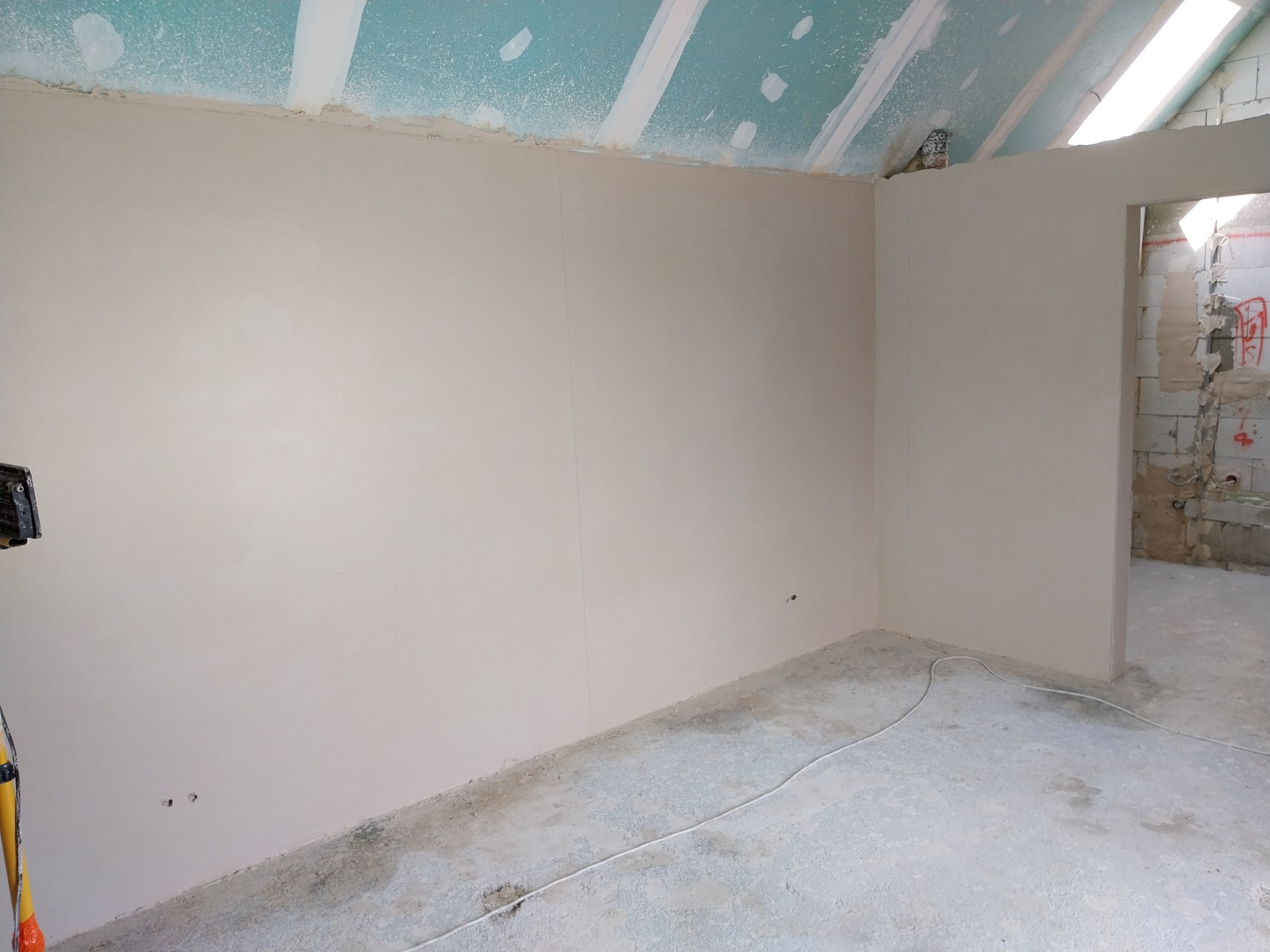 Wnętrze w trakcie prac wykończeniowych: gładko otynkowane ściany w jasnym odcieniu, sufit z płyt gipsowo-kartonowych, widoczne przewody elektryczne na podłodze, surowy stan pomieszczenia z widoczną...