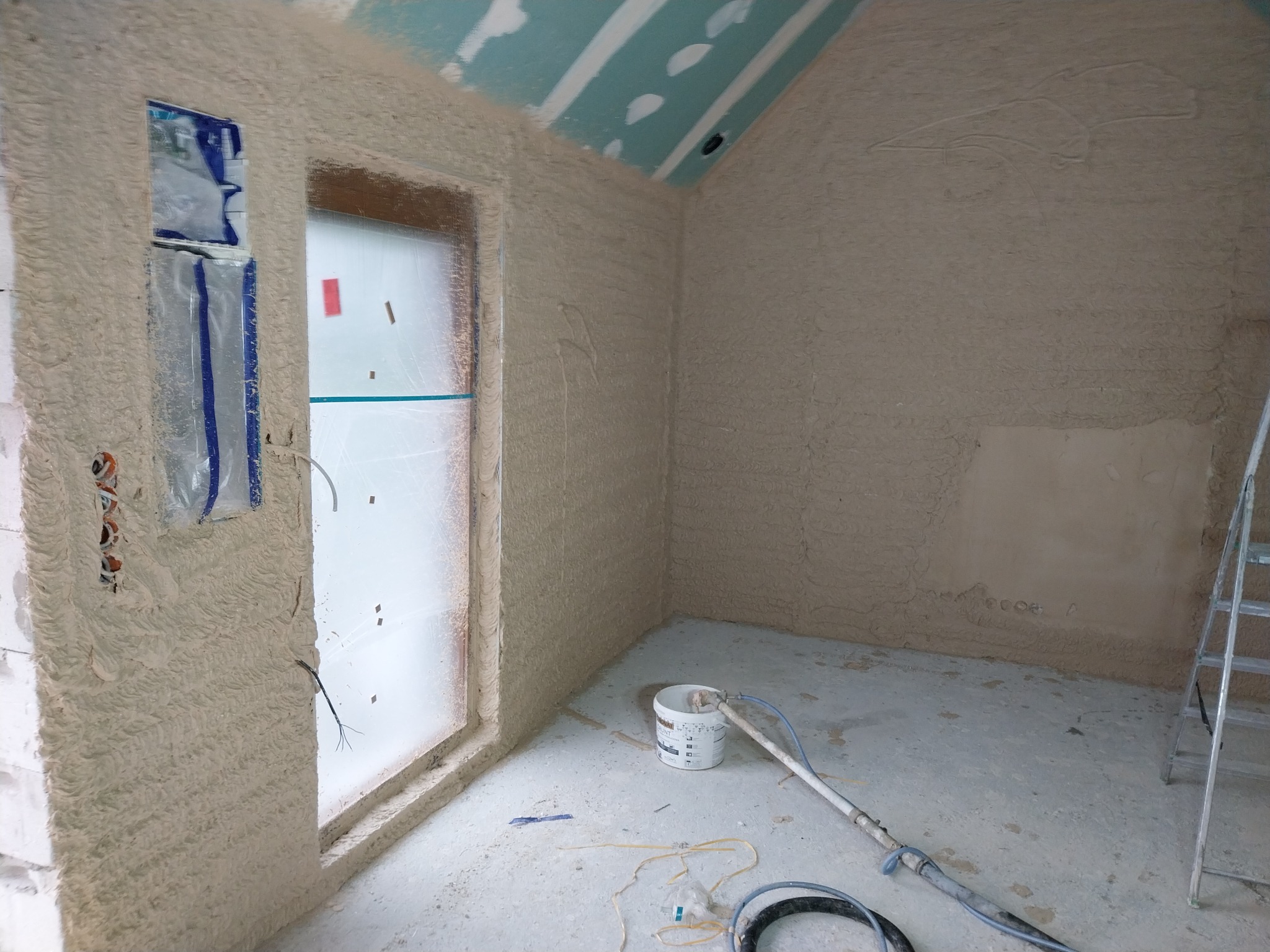 Wnętrze w trakcie tynkowania maszynowego, widoczne ściany pokryte świeżym tynkiem, okno zabezpieczone folią, oraz sprzęt do tynkowania i drabina