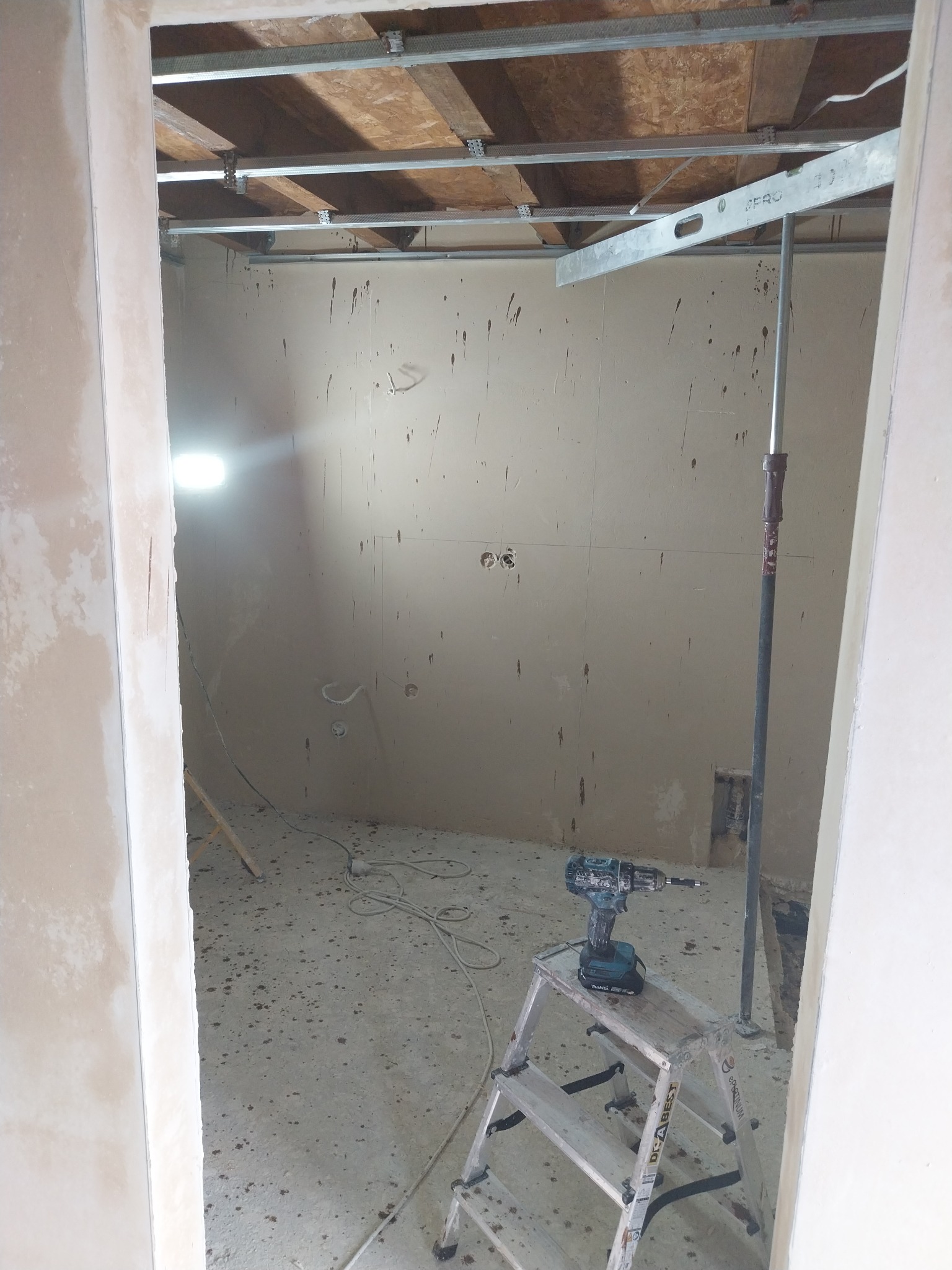 Wnętrze w trakcie prac wykończeniowych: ściany z płyt gipsowo-kartonowych z widocznymi śladami po szpachlowaniu, metalowa konstrukcja podwieszanego sufitu, poziomica oparta na rozpórce teleskopowej...