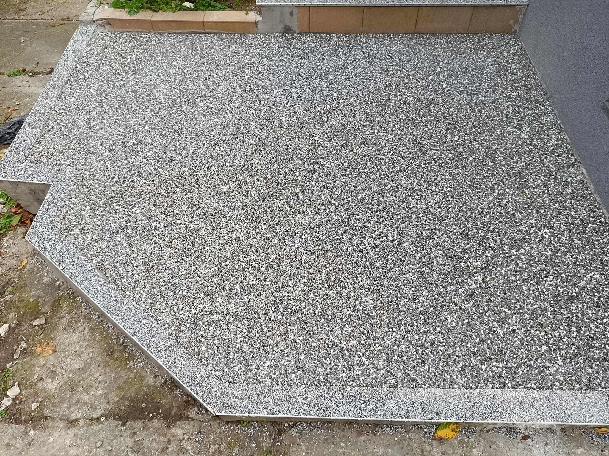 Wykończony taras z szarego, drobnoziarnistego kamienia żywicznego, obramowany metalową listwą, przylegający do ściany budynku i schodów.