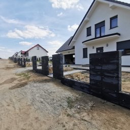 Arkadiusz Nowicki - Wznoszenie ogrodzenia z ciemnych, betonowych bloczków modułowych na posesji z nowoczesnym domem w tle, widoczne przygotowane fundamenty i narzędzia.