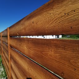 Arkadiusz Nowicki - Drewniane ogrodzenie poziome, zabezpieczone lakierem, z widocznymi kroplami wody, na tle błękitnego nieba i fragmentu białego budynku.