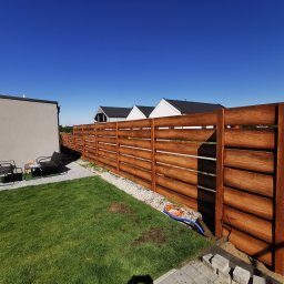 Arkadiusz Nowicki - Nowoczesne ogrodzenie drewniane w Śremie z poziomych desek o wyraźnym usłojeniu, oddzielające posesję z trawnikiem i zestawem mebli ogrodowych od sąsiednich domów pod błękitnym niebem.