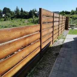 Arkadiusz Nowicki - Długie, poziome przęsła ogrodzenia z imitacją słojów drewna, oddzielające trawnik od wyłożonego kostką brukową podwórka, z widocznym w tle zielonym krajobrazem.