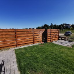 Arkadiusz Nowicki - Nowoczesne, poziome brązowe panele ogrodzeniowe z imitacją drewna oddzielające trawnik od posesji z samochodem, widoczne kostki brukowe i przygotowane podłoże.