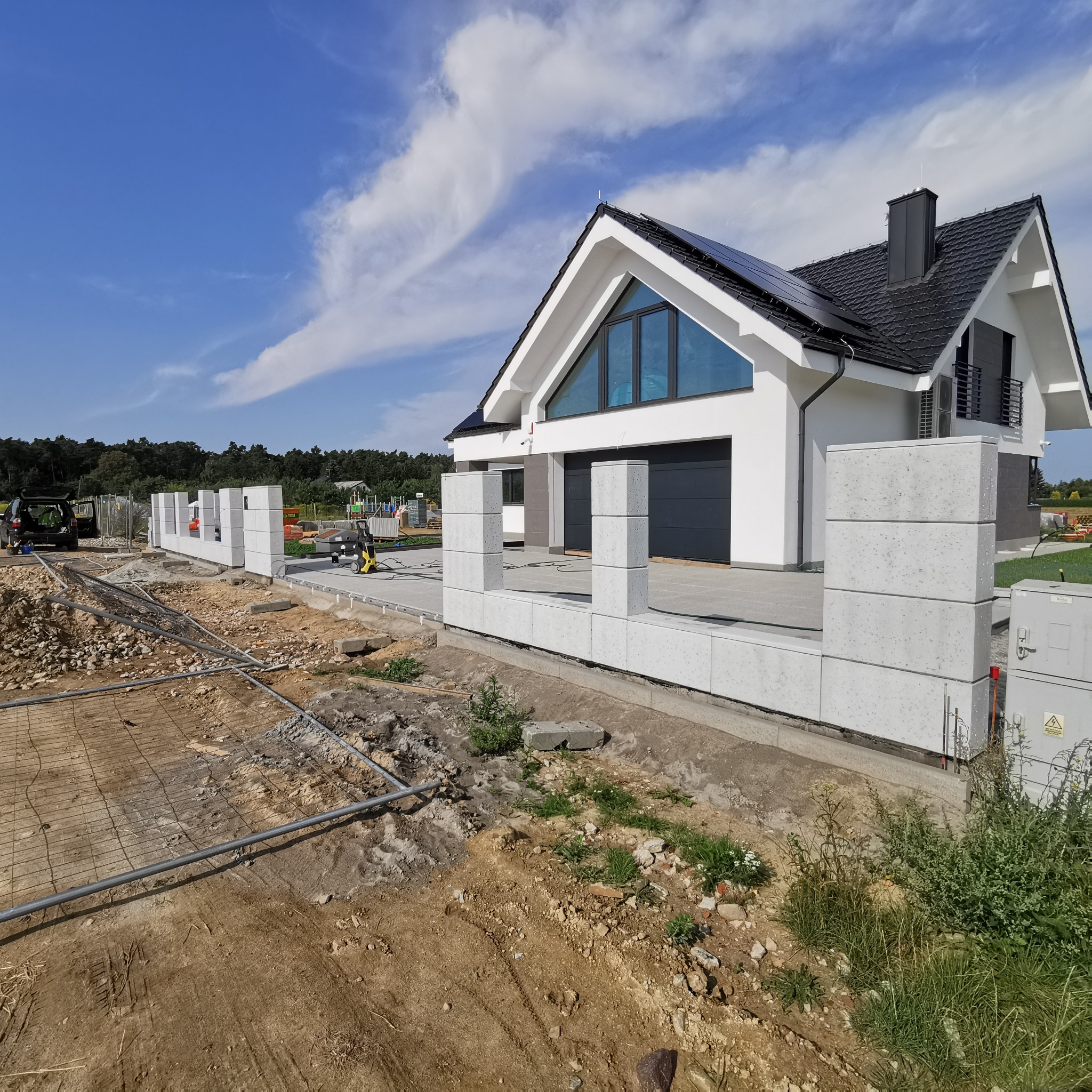 Montaż ogrodzenia z prefabrykatów betonowych na tle nowoczesnego domu z panelami słonecznymi i błękitnym niebem.