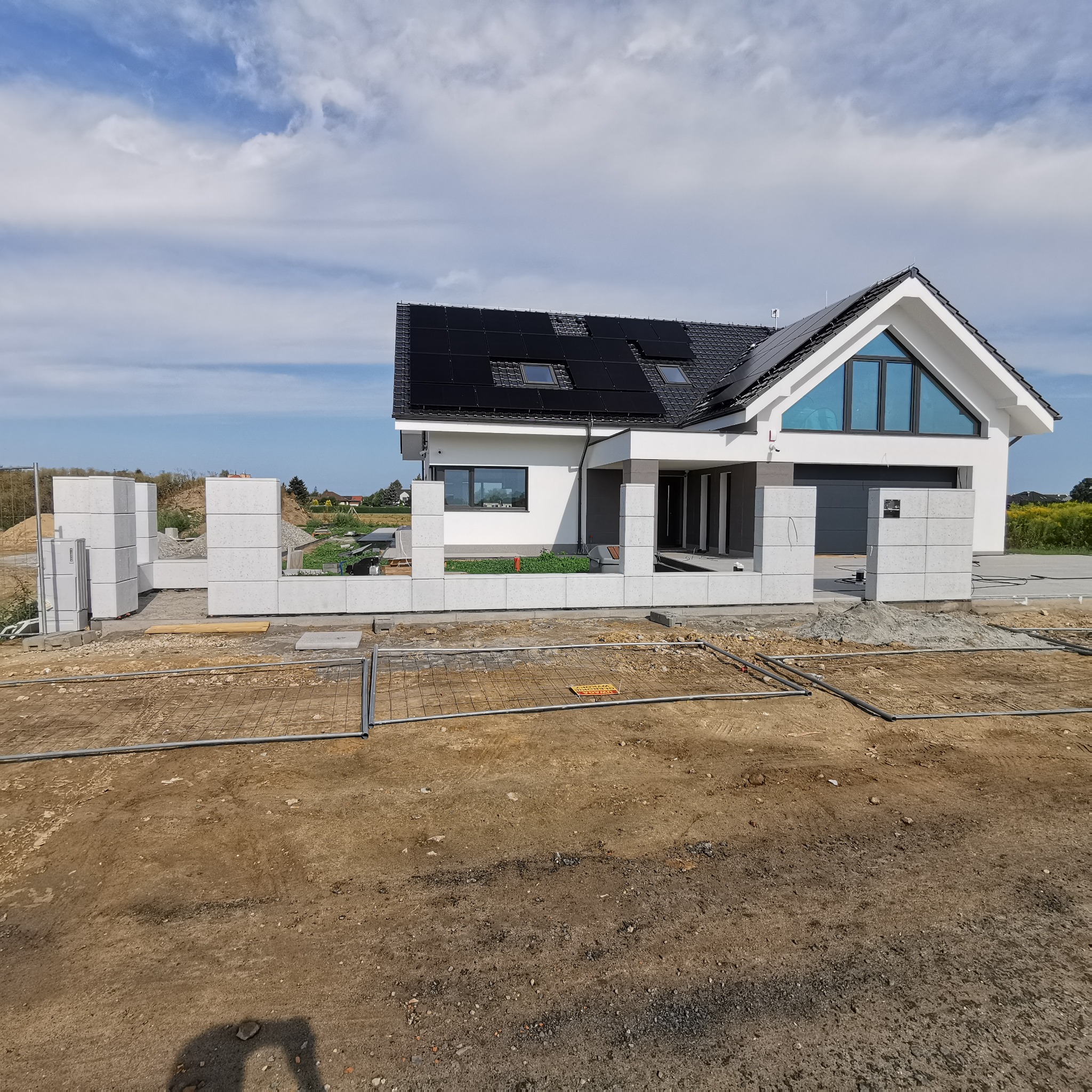 Wznoszona konstrukcja z prefabrykatów betonowych przed nowoczesnym domem z panelami słonecznymi na dachu, widoczne zbrojenie w ziemi i elementy instalacji.