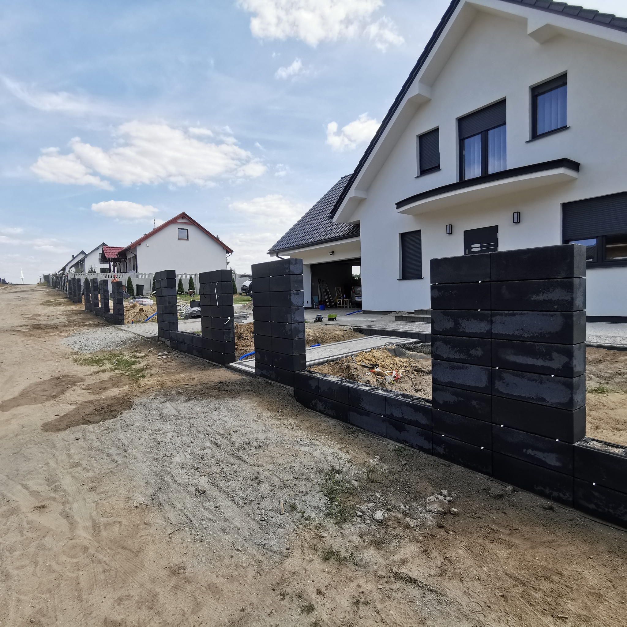 Wznoszenie ogrodzenia z ciemnych, betonowych bloczków modułowych na posesji z nowoczesnym domem w tle, widoczne przygotowane fundamenty i narzędzia.