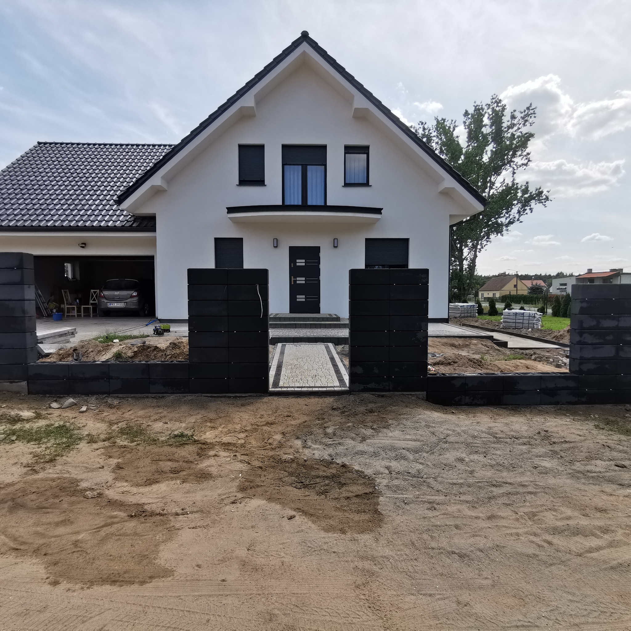 Nowoczesny dom jednorodzinny z czarnym dachem i białymi ścianami, w trakcie budowy ogrodzenia z betonowych bloczków. Widoczny fragment chodnika z kostki prowadzącego do wejścia. W tle garaż...