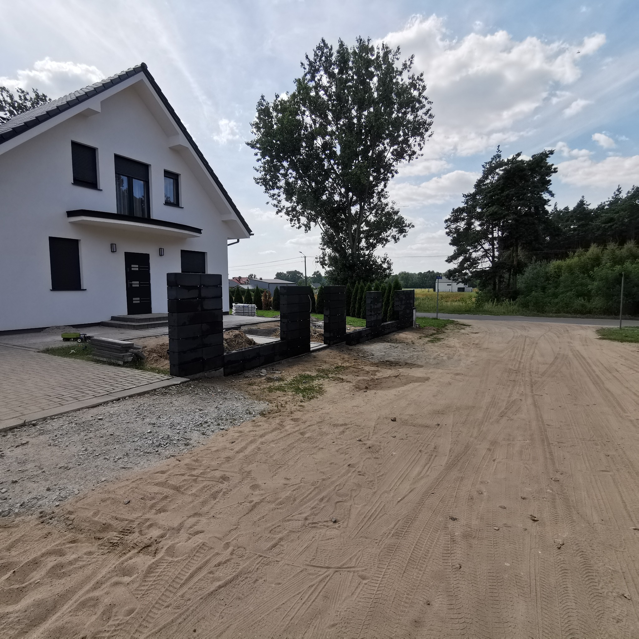 Fragment czarnego ogrodzenia z prefabrykatów betonowych w trakcie montażu przed nowoczesnym, białym domem z czarnymi oknami i drzwiami, widoczny plac budowy i piasek.