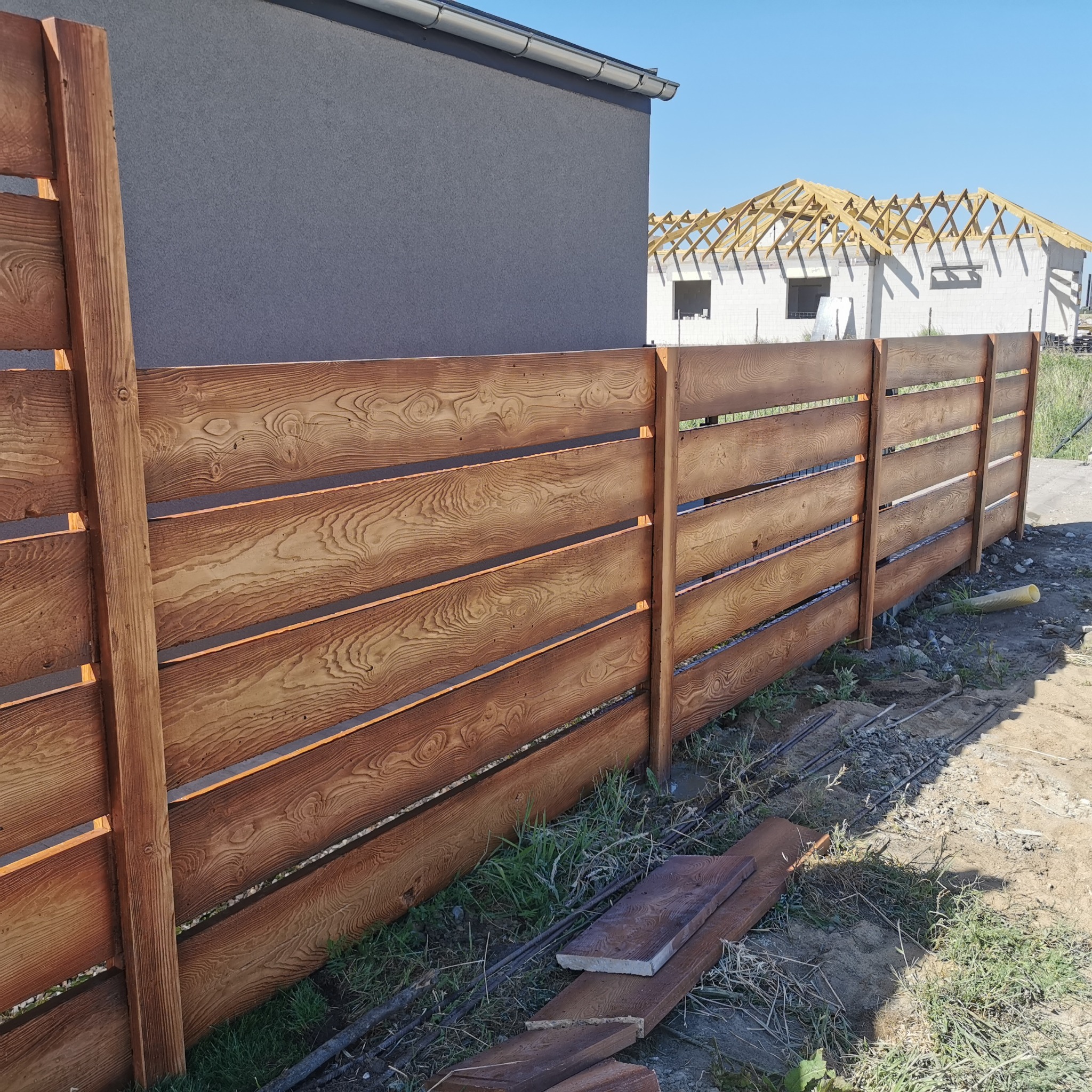 Nowoczesne ogrodzenie poziome z brązowych desek z wyraźnym rysunkiem słojów drewna, na tle szarego domu i budowy w stanie surowym otwartym.