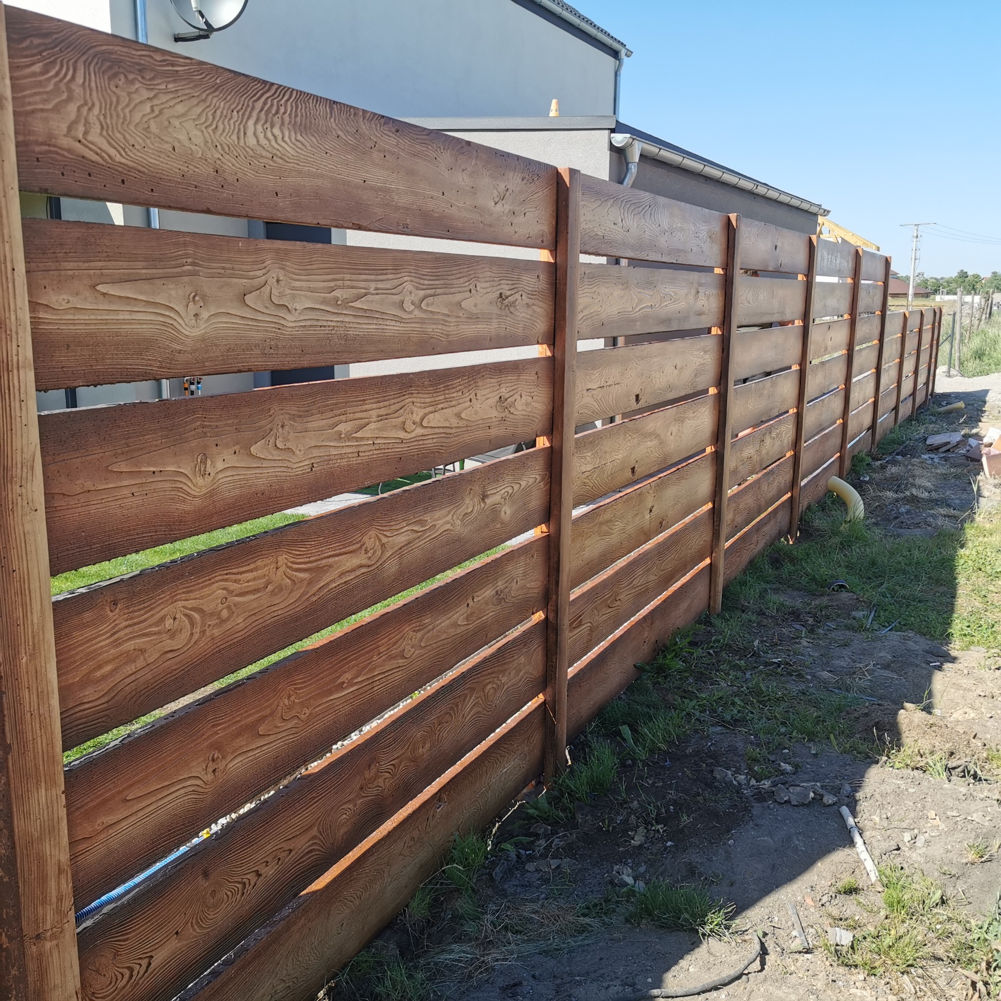 Długi, poziomy płot z brązowych desek z widocznym rysunkiem słojów drewna, oddzielający posesję od drogi, z fragmentem domu w tle.