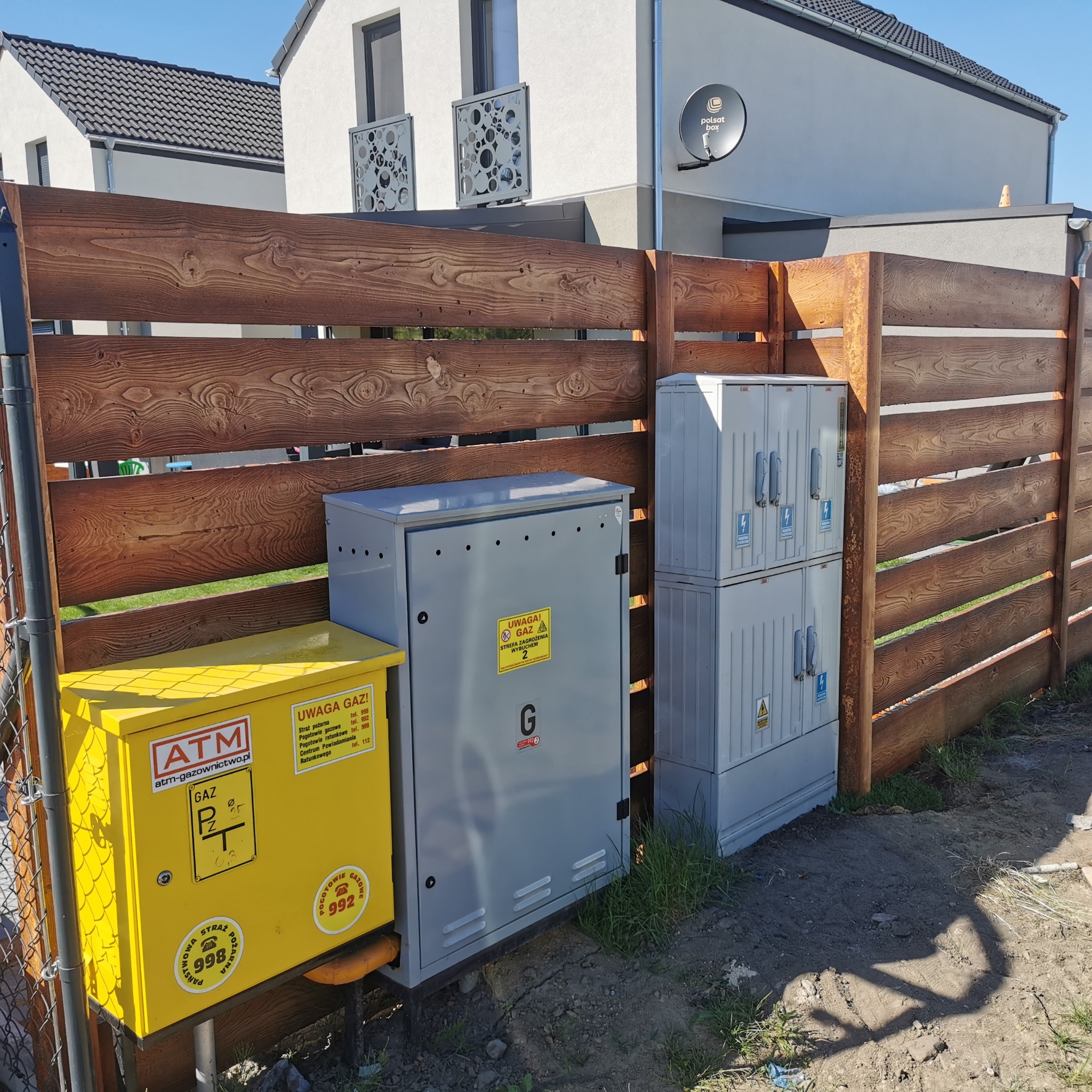 Drewniane ogrodzenie z brązowych desek, za nim skrzynki gazowe i elektryczne w kolorach żółtym i szarym, na tle domy jednorodzinne.