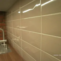 Beżowe, błyszczące płytki ceramiczne na ścianie, z widoczną fugą, fragment ściany z cegły i metalowa bateria umywalkowa.