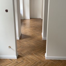 F.H.U Michał Gawor docieplenia budynków - Wnętrze mieszkania z ułożoną podłogą z płytek imitujących drewno w jodełkę, widok na kolejne pomieszczenia przez symetryczne przejścia w ścianach, ściany pomalowane na jasny odcień.