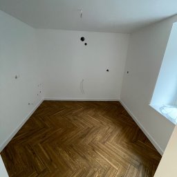 F.H.U Michał Gawor docieplenia budynków - Pusty pokój z parkietem ułożonym w jodełkę, białe ściany z widocznymi otworami na instalacje elektryczne i wystającym kablem, marmurowy parapet okienny.