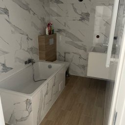 F.H.U Michał Gawor docieplenia budynków - Nowoczesna łazienka z marmurowymi płytkami na ścianach i obudowie wanny, drewnopodobne płytki na podłodze, wisząca szafka z umywalką i toaleta.