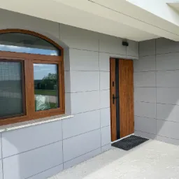 Nowoczesna elewacja budynku z szarymi panelami, brązowymi drzwiami i oknem z łukiem, widoczny fragment surowego, betonowego podłoża.