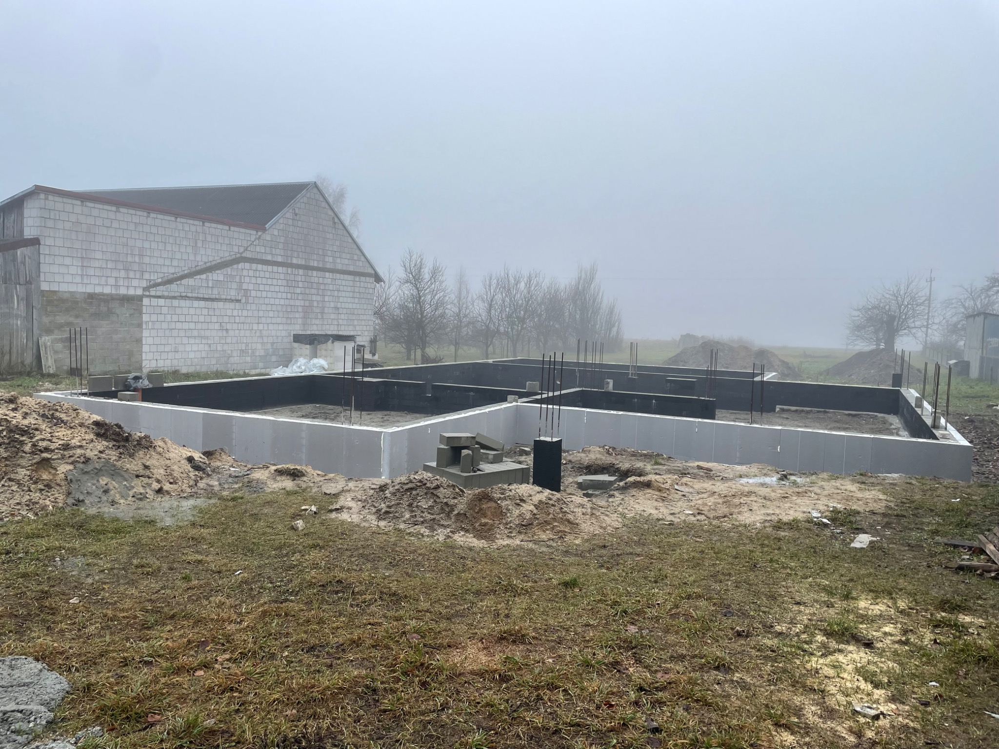 Fundament domu w trakcie budowy, ocieplony styropianem, widoczne zbrojenia, w tle budynek gospodarczy i mglista pogoda.