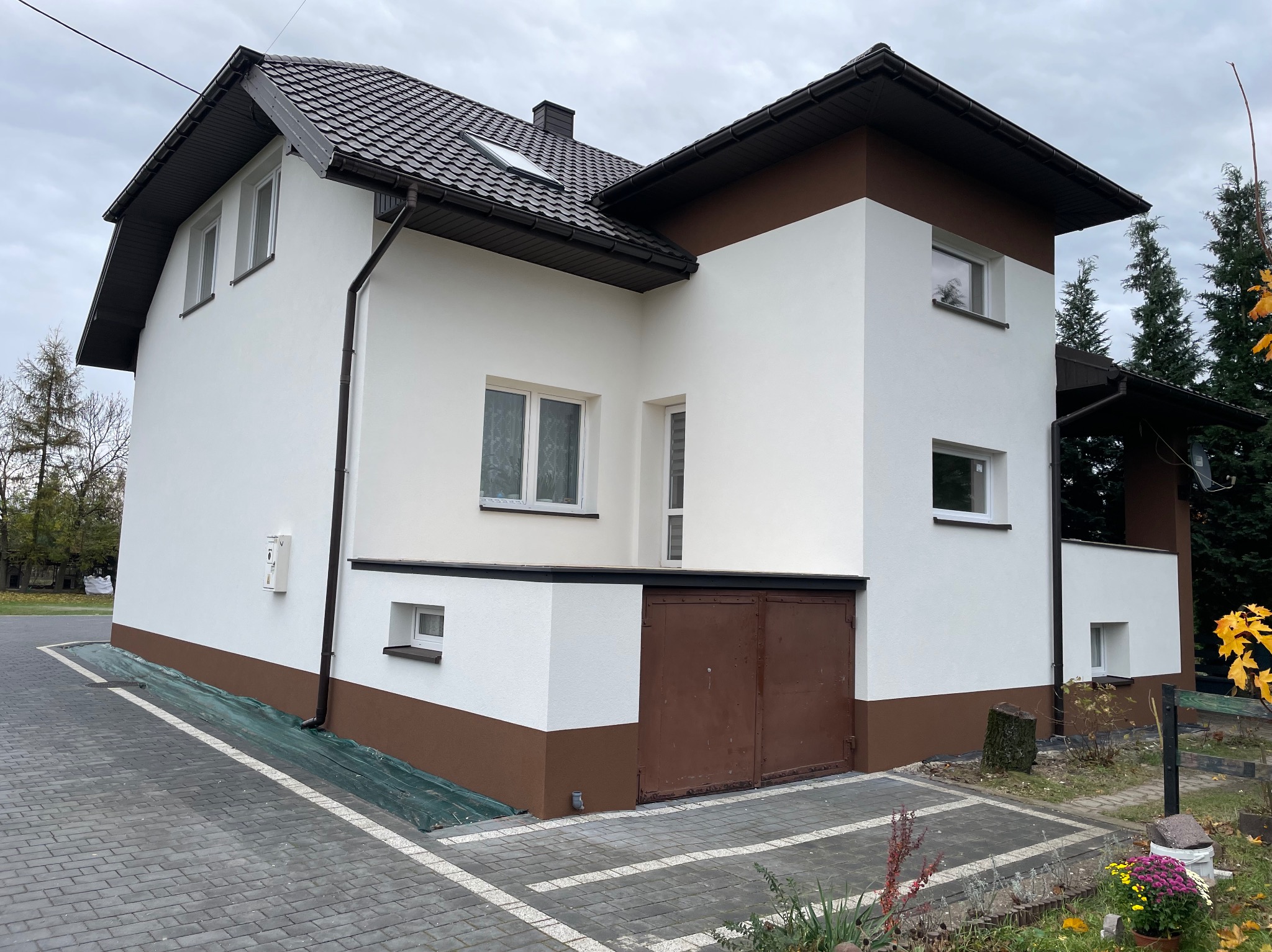 Dwukondygnacyjny dom z białą elewacją, brązowymi akcentami i brązowym dachem, widoczny z poziomu chodnika z kostki brukowej, z zieloną plandeką zabezpieczającą przy podstawie ściany.