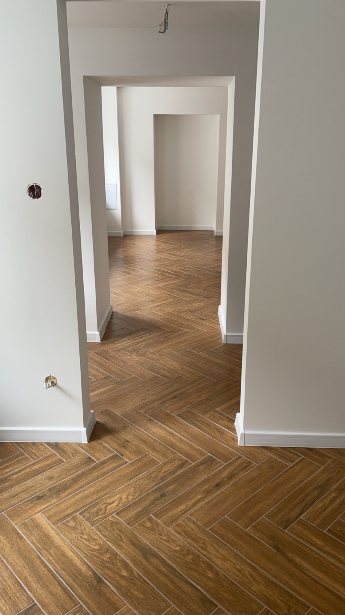 Wnętrze mieszkania z ułożoną podłogą z płytek imitujących drewno w jodełkę, widok na kolejne pomieszczenia przez symetryczne przejścia w ścianach, ściany pomalowane na jasny odcień.