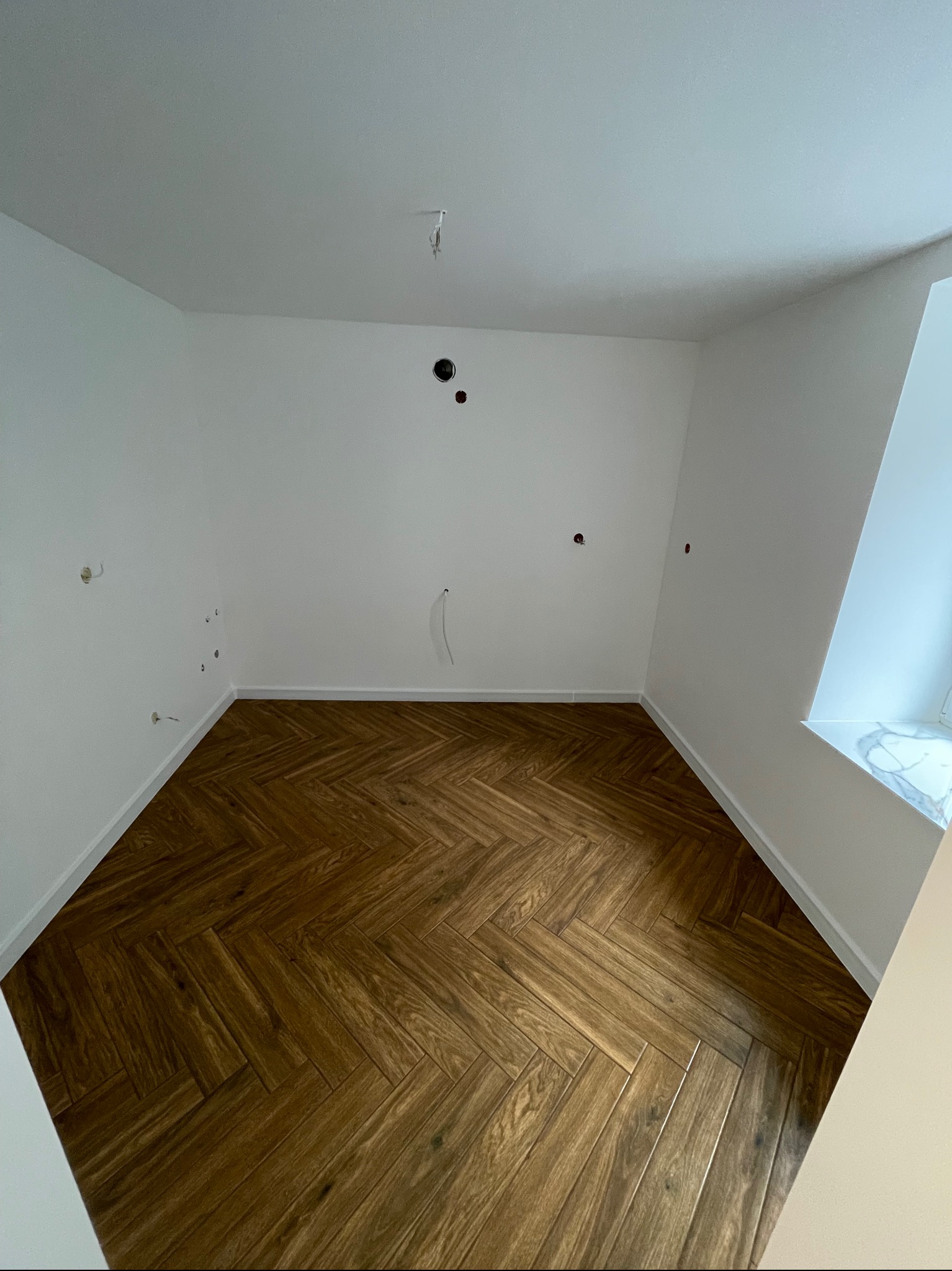 Pusty pokój z parkietem ułożonym w jodełkę, białe ściany z widocznymi otworami na instalacje elektryczne i wystającym kablem, marmurowy parapet okienny.