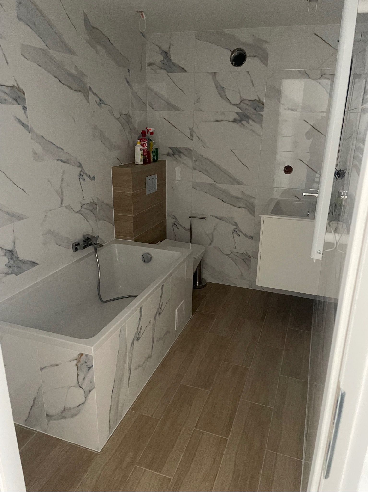 Nowoczesna łazienka z marmurowymi płytkami na ścianach i obudowie wanny, drewnopodobne płytki na podłodze, wisząca szafka z umywalką i toaleta.