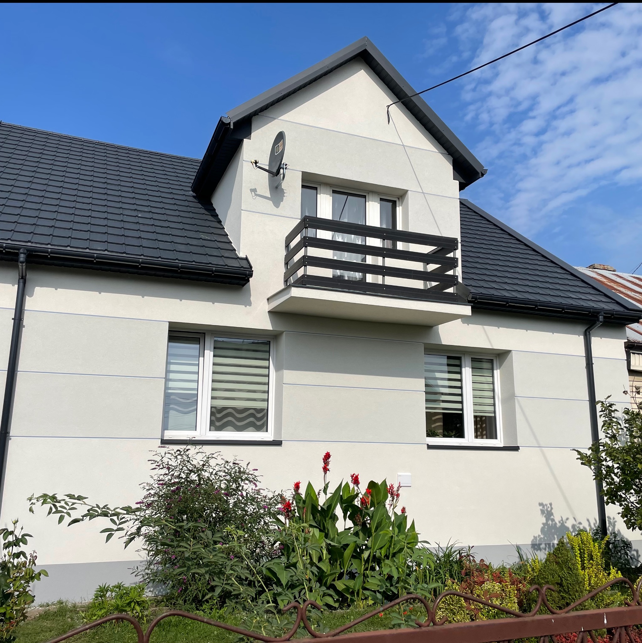 Jasnoszary dom jednorodzinny z ciemną dachówką, balkonem z czarną balustradą i anteną satelitarną na elewacji, otoczony zielenią i kwiatami w słoneczny dzień.