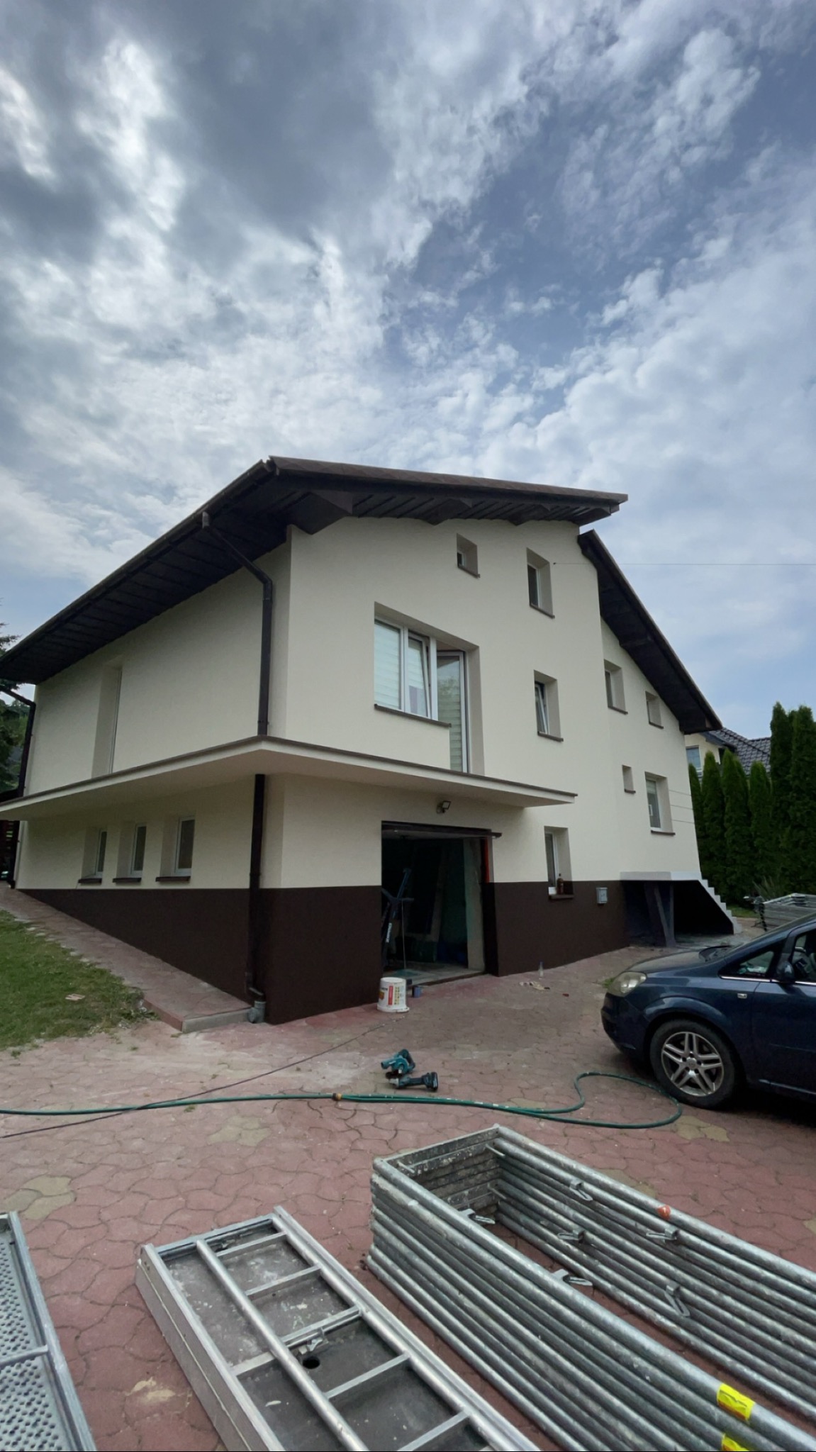 Dwukondygnacyjny dom po częściowym odnowieniu elewacji, z kremową górną częścią i brązową dolną, widoczne rusztowania, narzędzia i zaparkowany samochód.