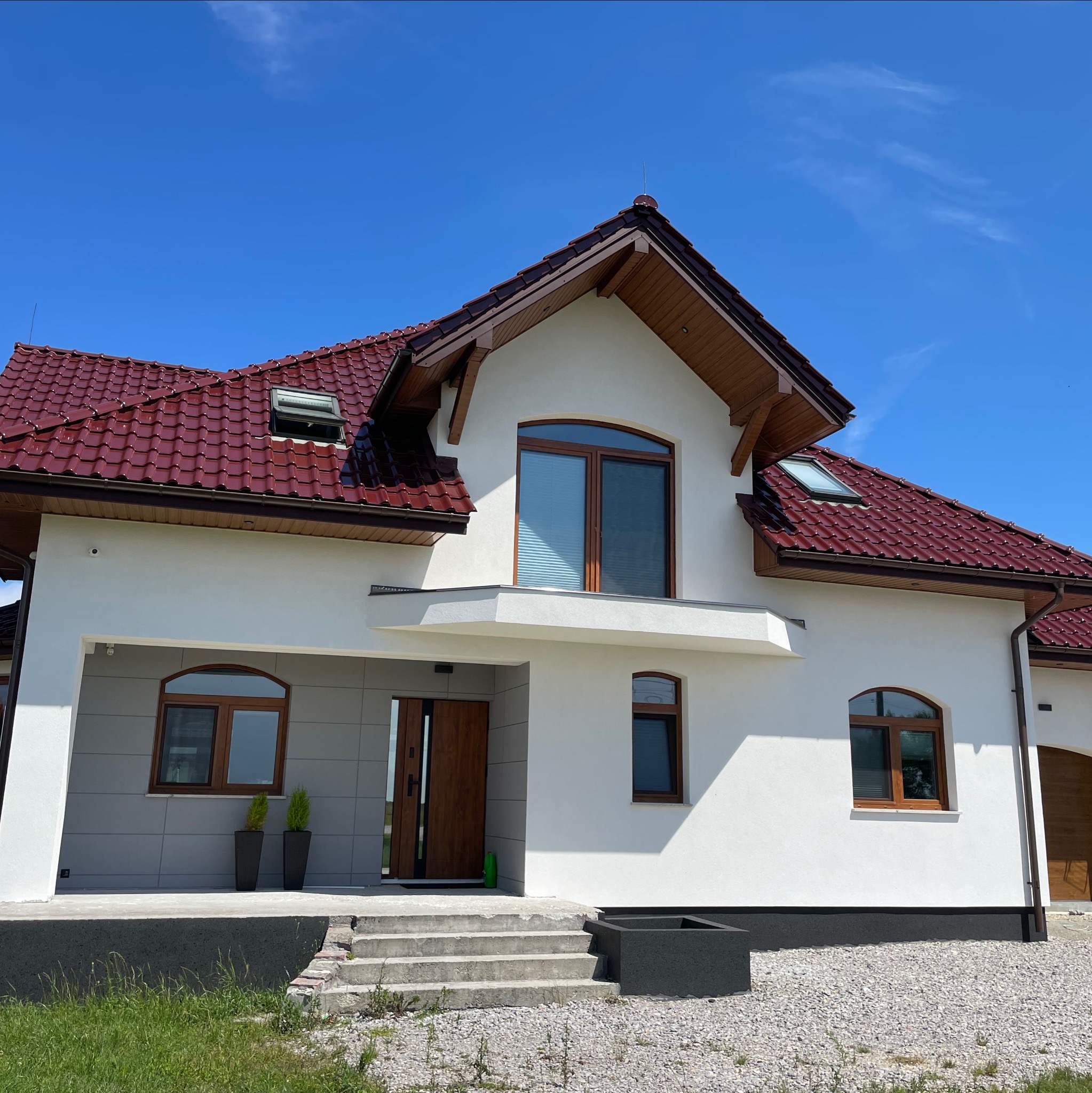 Nowoczesny dom jednorodzinny z białą elewacją, czerwoną dachówką i drewnianymi oknami w słoneczny dzień. Widoczne schody wejściowe, zadbany trawnik i błękitne niebo.