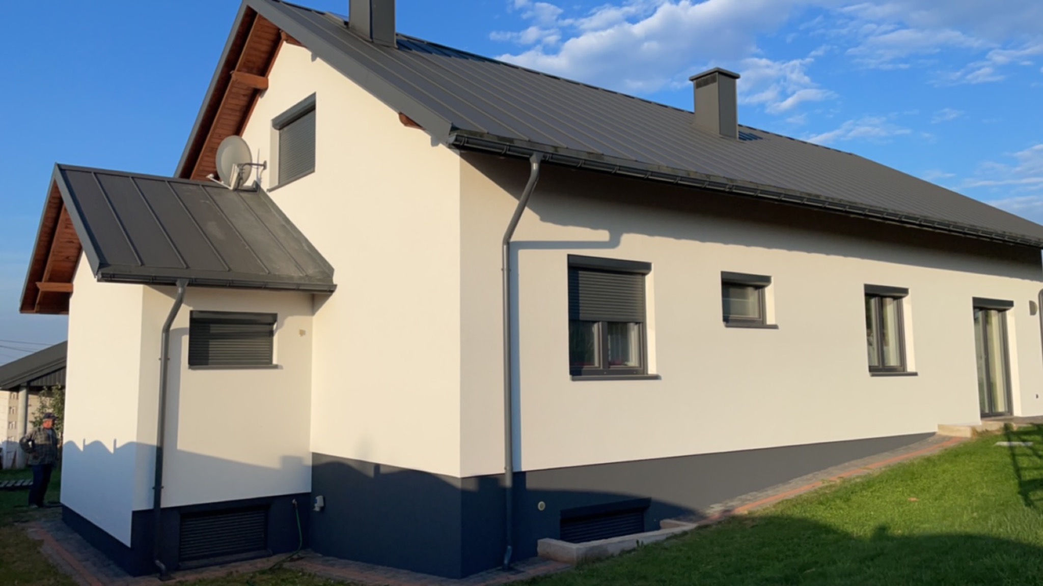 Nowoczesny dom jednorodzinny z jasną elewacją, szarą podmurówką i ciemnoszarym dachem z blachy na tle błękitnego nieba.