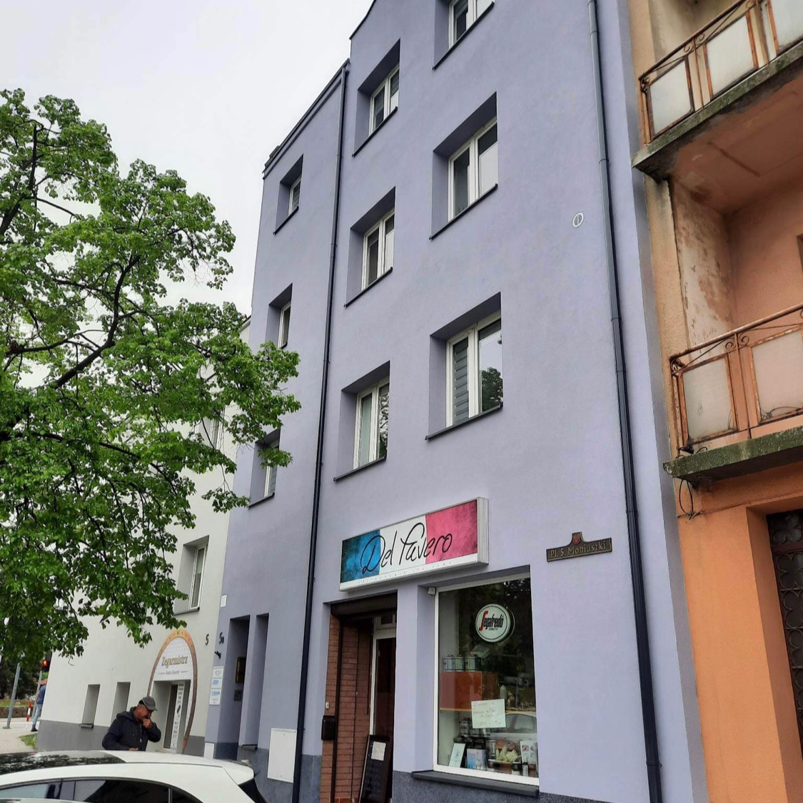 Odnowiona elewacja kamienicy w odcieniach szarości z nowymi oknami, widok z dołu, obok drzewo i sąsiedni budynek z balkonami.