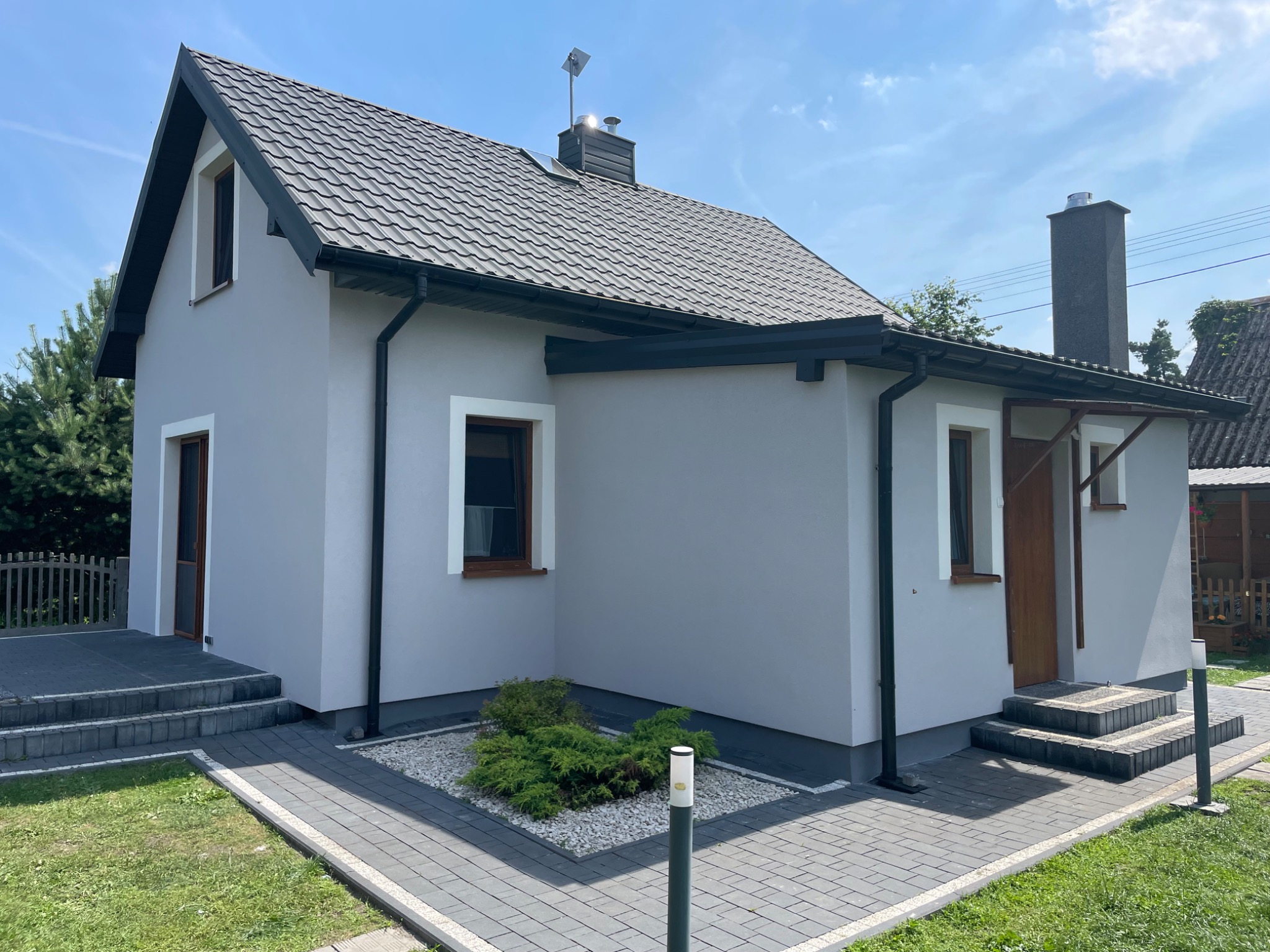 Dom jednorodzinny z szarą elewacją, ciemną dachówką i drewnianą stolarką okienną, otoczony trawnikiem i kostką brukową.