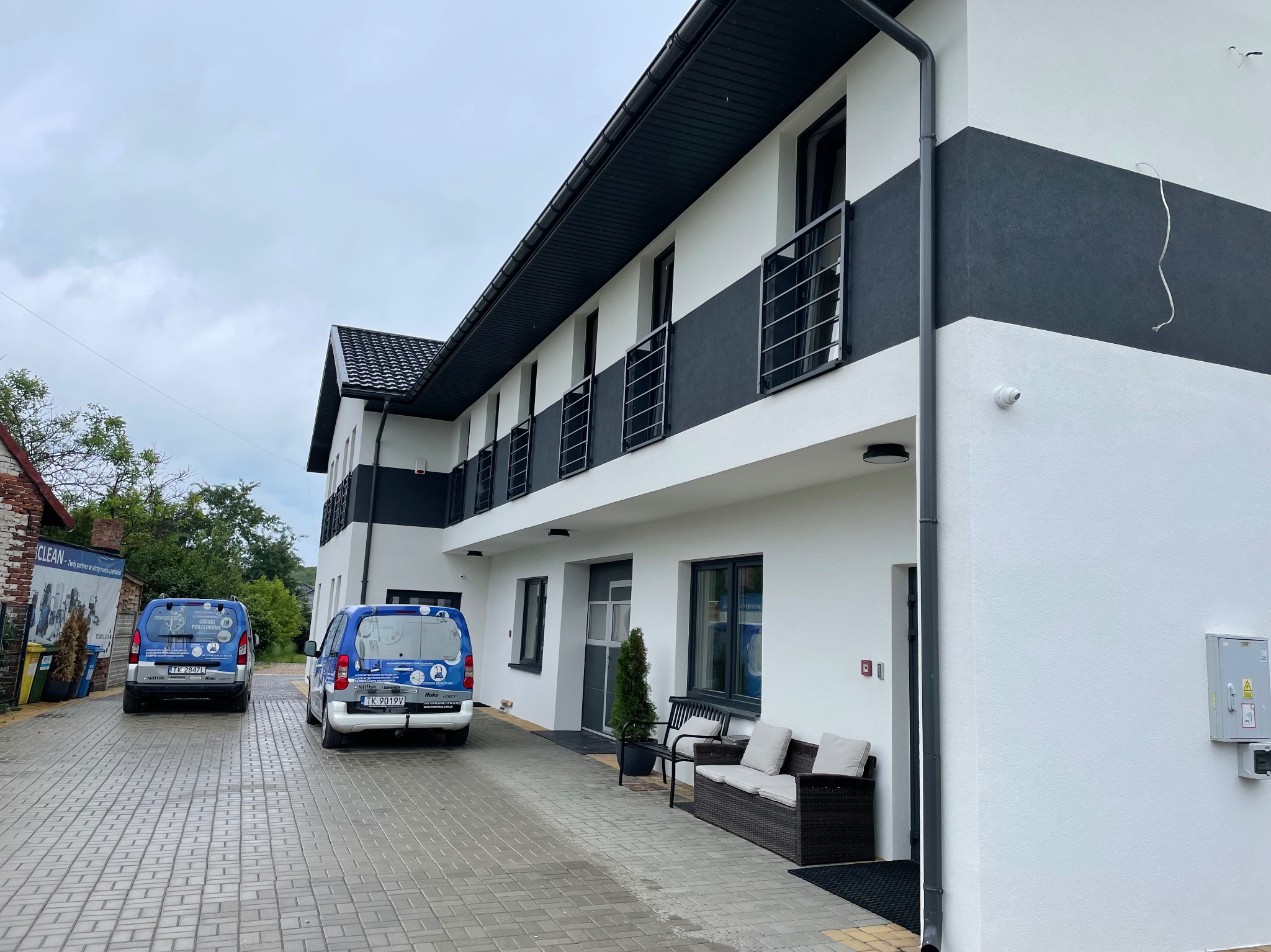 Nowoczesny budynek po renowacji elewacji, z dwoma samochodami dostawczymi firmy sprzątającej zaparkowanymi przed wejściem.