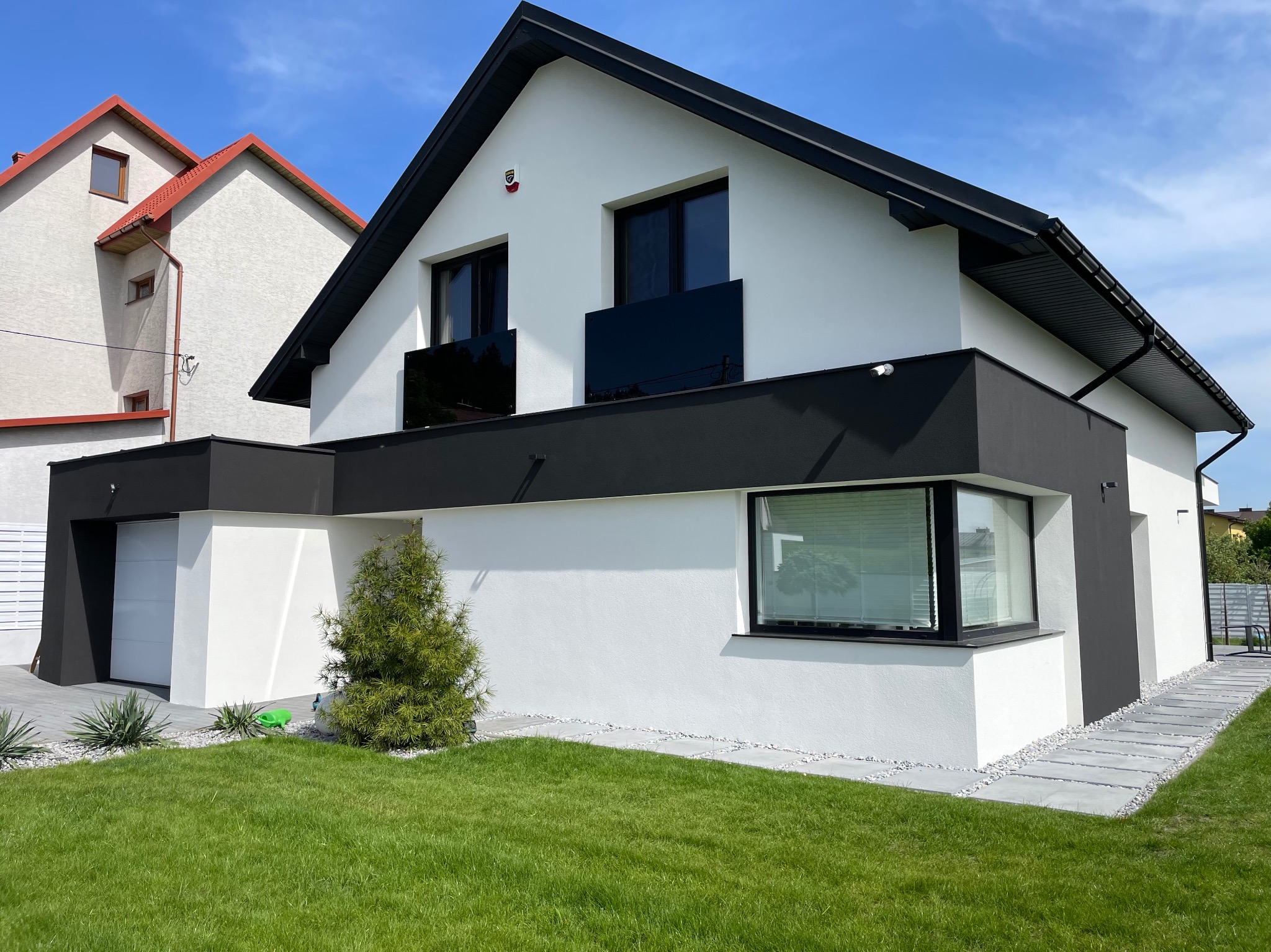 Nowoczesny dom jednorodzinny z białą elewacją i czarnymi akcentami, widok z przodu na zadbany trawnik i elementy dekoracyjne z kamienia.