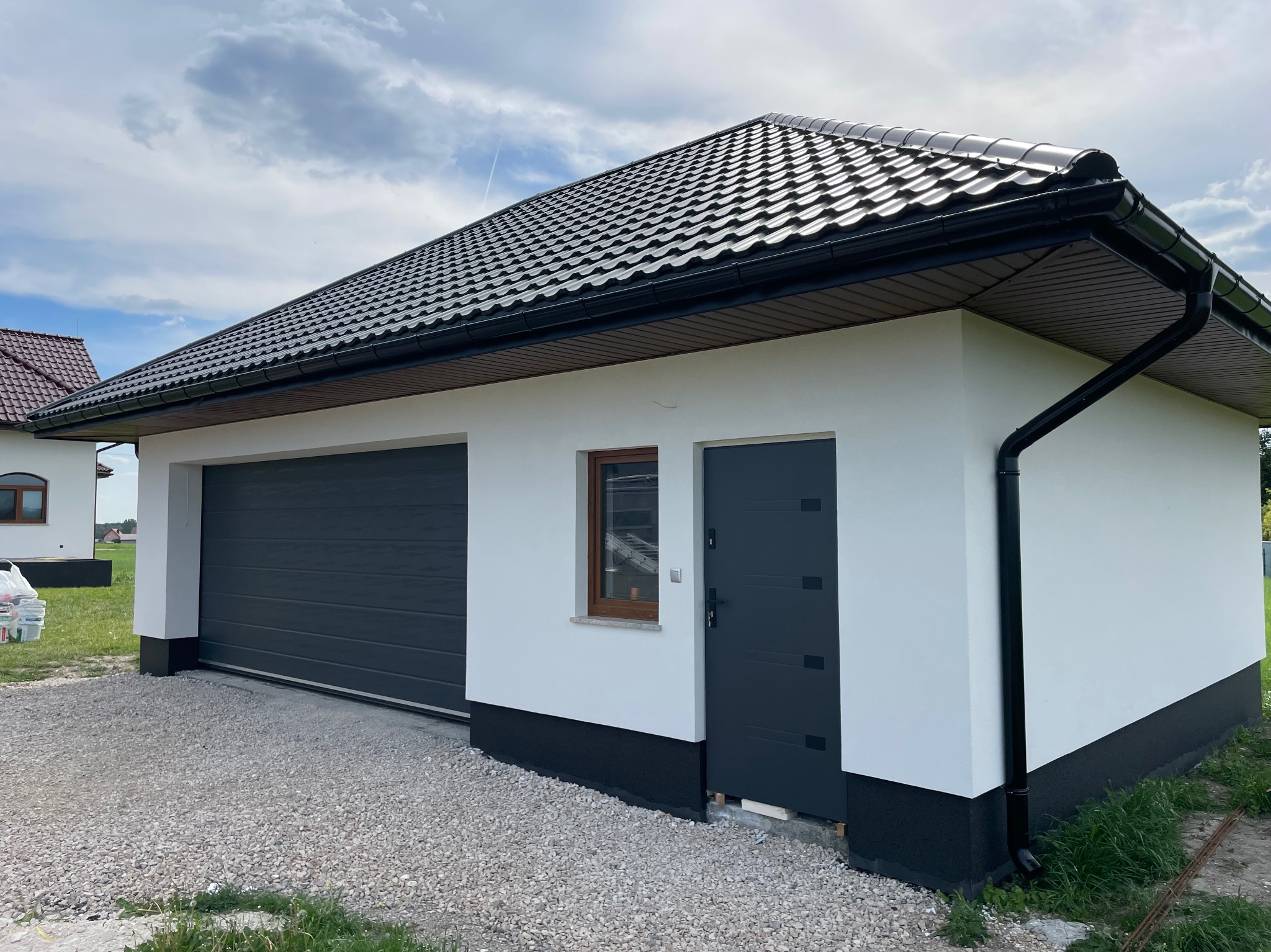 Nowoczesny budynek gospodarczy z białą elewacją, grafitową blachodachówką i bramą garażową, widok na świeżo położony tynk i obróbki blacharskie.