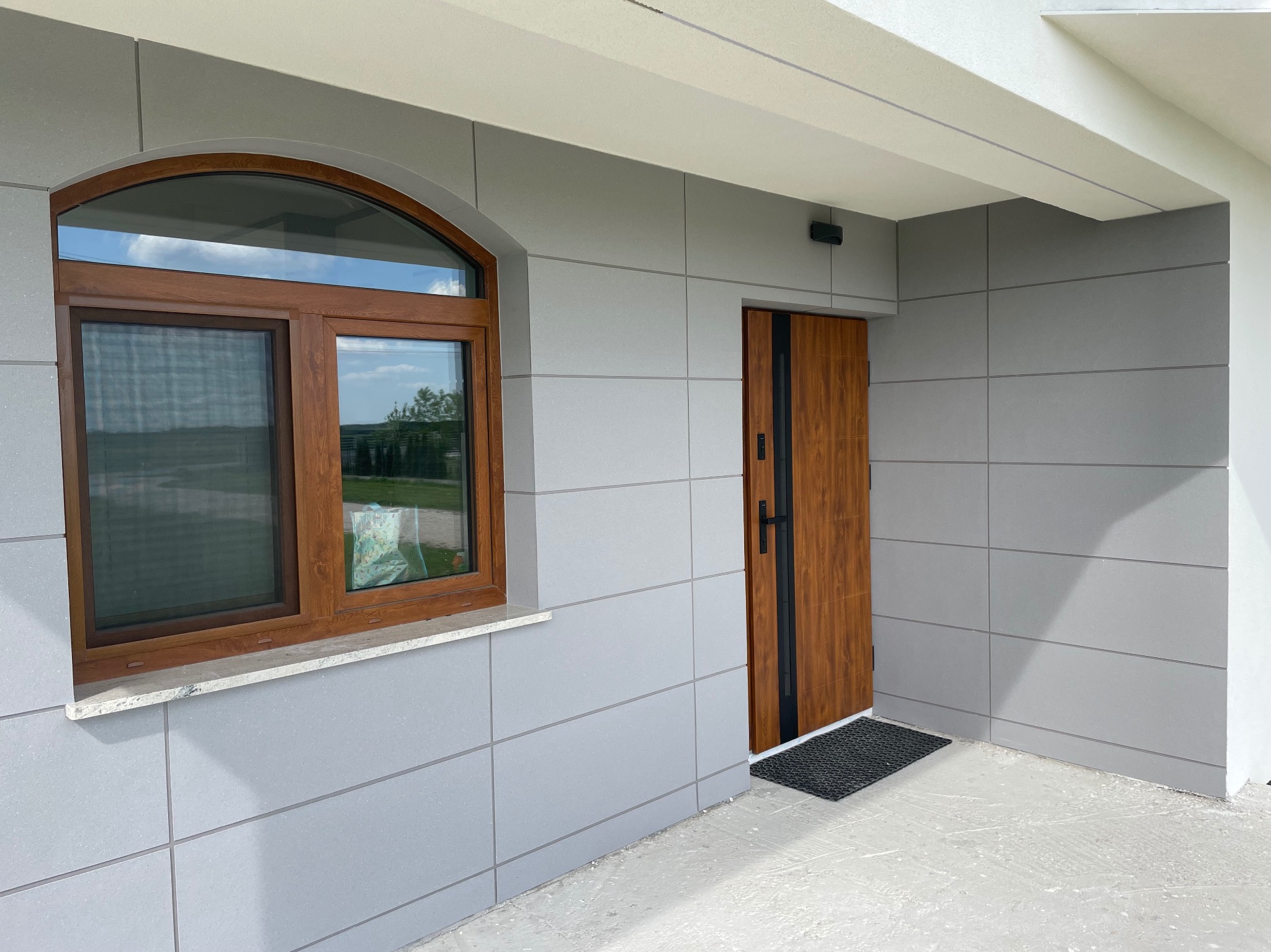 Nowoczesna elewacja budynku z szarymi panelami, brązowymi drzwiami i oknem z łukiem, widoczny fragment surowego, betonowego podłoża.