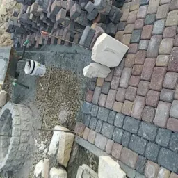 Wykonywanie nawierzchni z kostki brukowej o nieregularnych kształtach i kolorach, widoczne przygotowanie terenu i materiały budowlane, w tle drzewa.