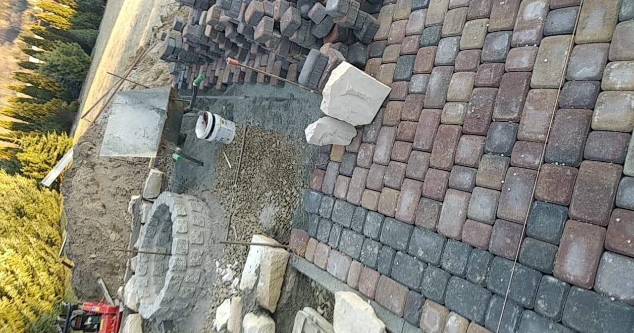 Wykonywanie nawierzchni z kostki brukowej o nieregularnych kształtach i kolorach, widoczne przygotowanie terenu i materiały budowlane, w tle drzewa.