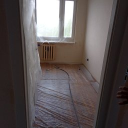 My House recovery - Pomieszczenie przygotowane do malowania: podłoga zabezpieczona folią, okno i grzejnik osłonięte, widoczny pistolet natryskowy z przewodem.